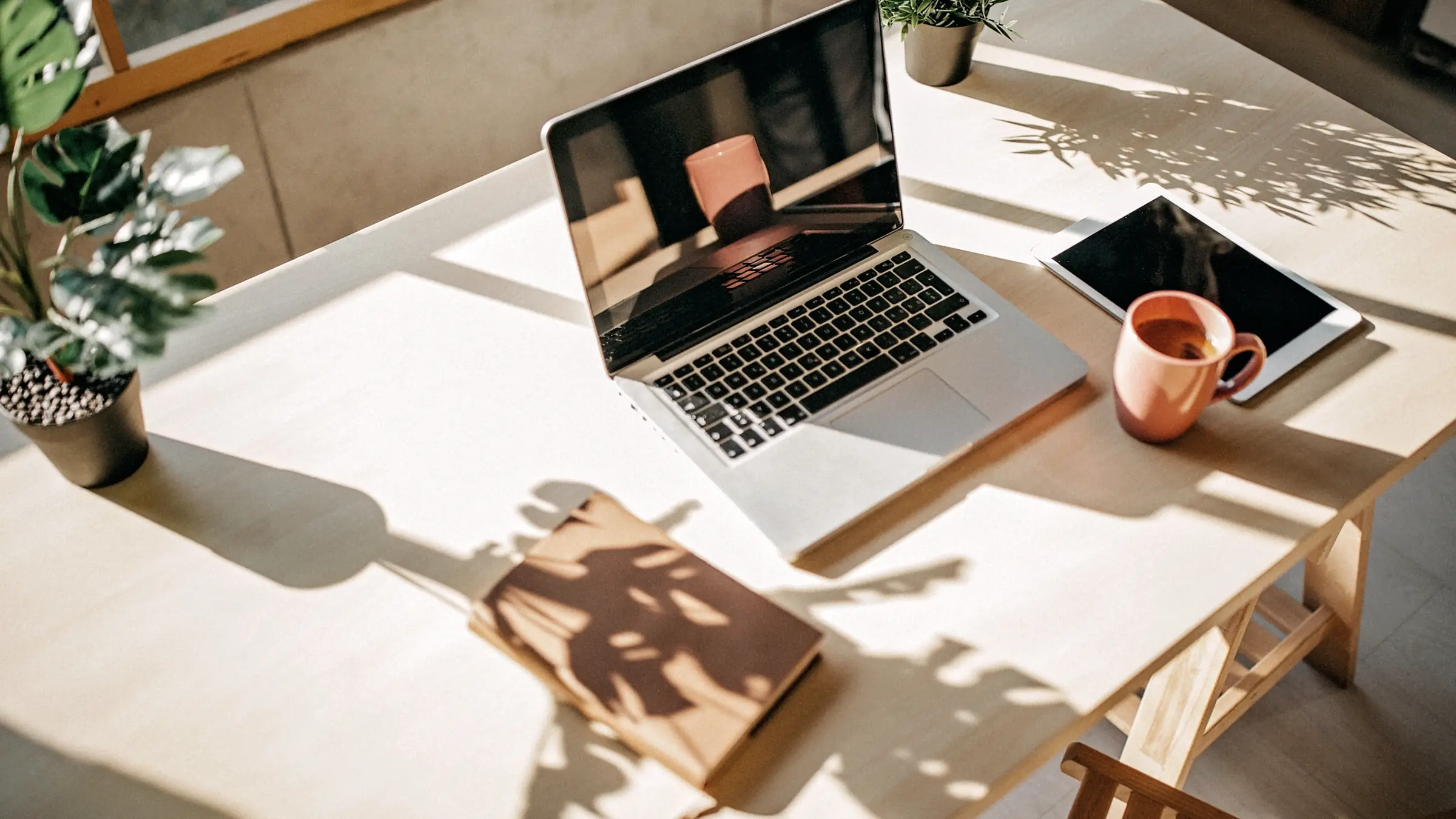 Heller Holzschreibtisch im Sonnenlicht mit Laptop, Tablet, Kaffeetasse und Notizbuch